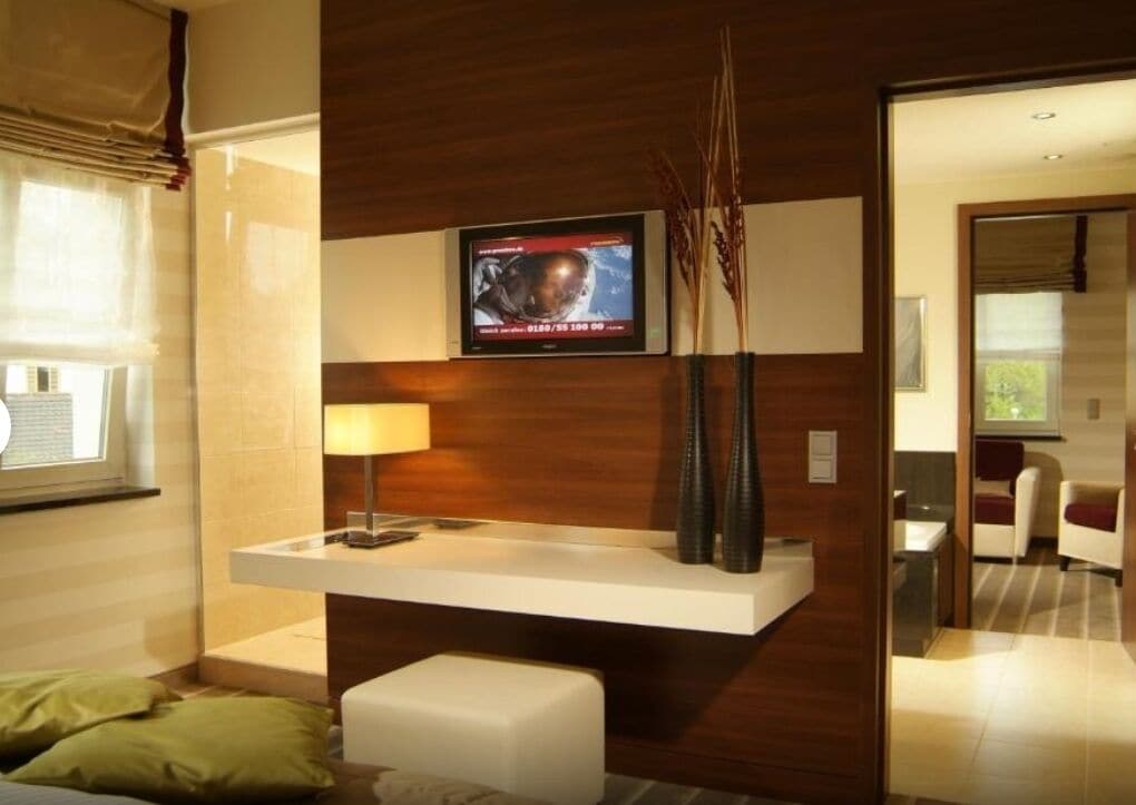 A modern hotel room with a wooden accent wall, a flat-screen TV mounted above a white console table, a bedside lamp, and a green pillow on the bed.