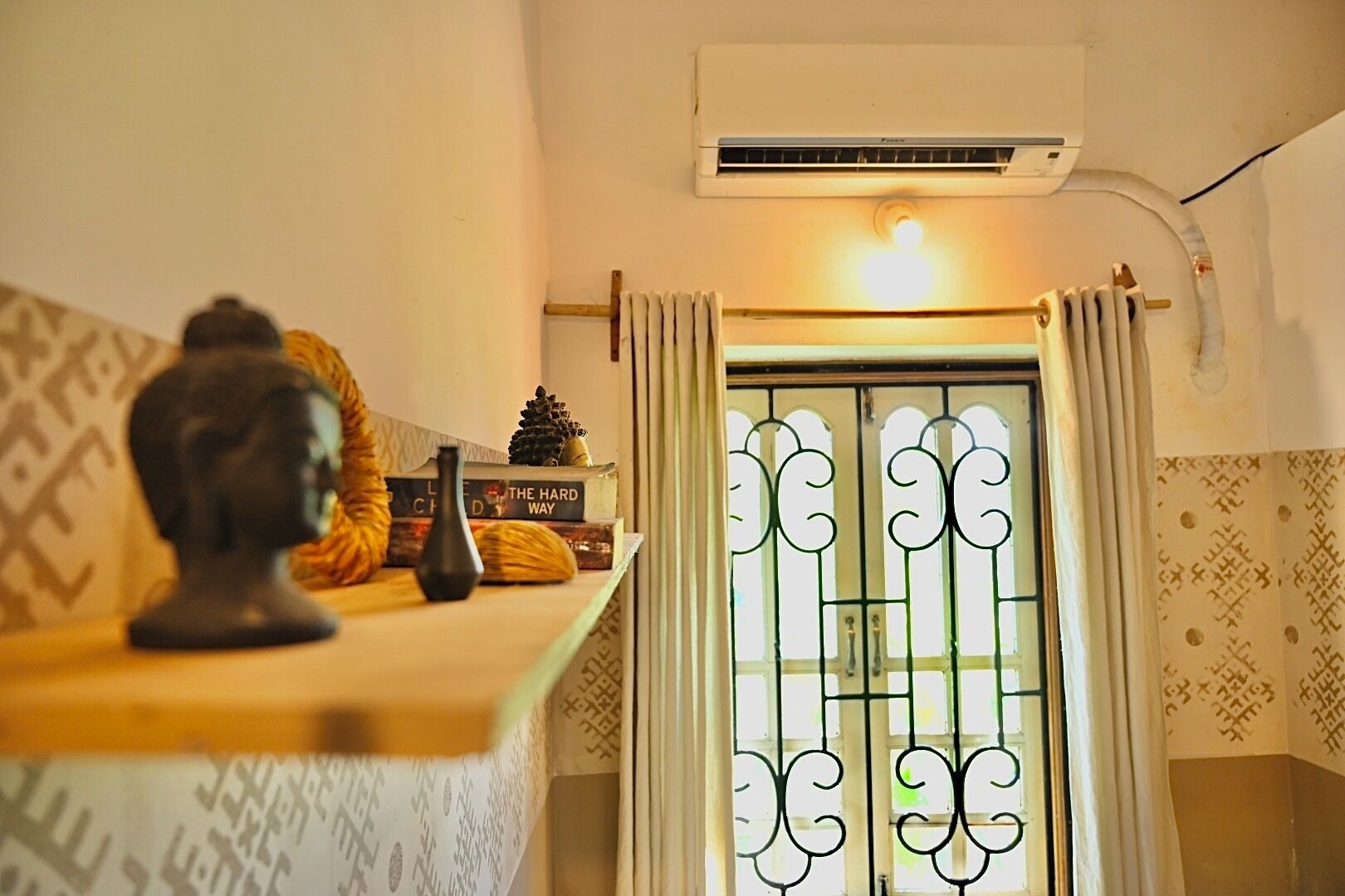 A room with a sofa, a small table with decorative items, a wall-mounted air conditioner, and a window with decorative iron bars.
