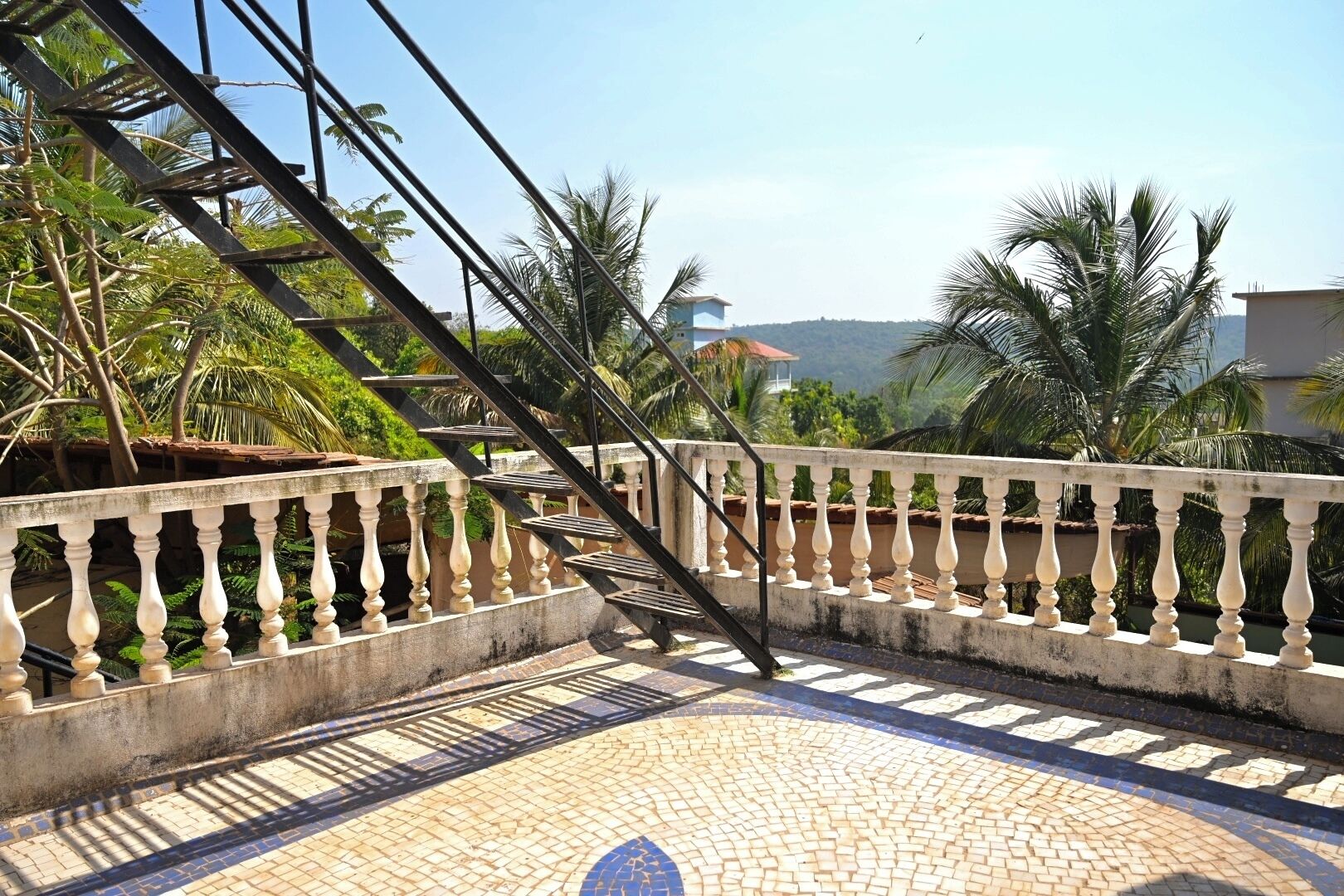Un escalier agrémenté d’une balustrade mène à un balcon qui offre une vue sur un paysage luxuriant, avec des palmiers et une colline lointaine.
