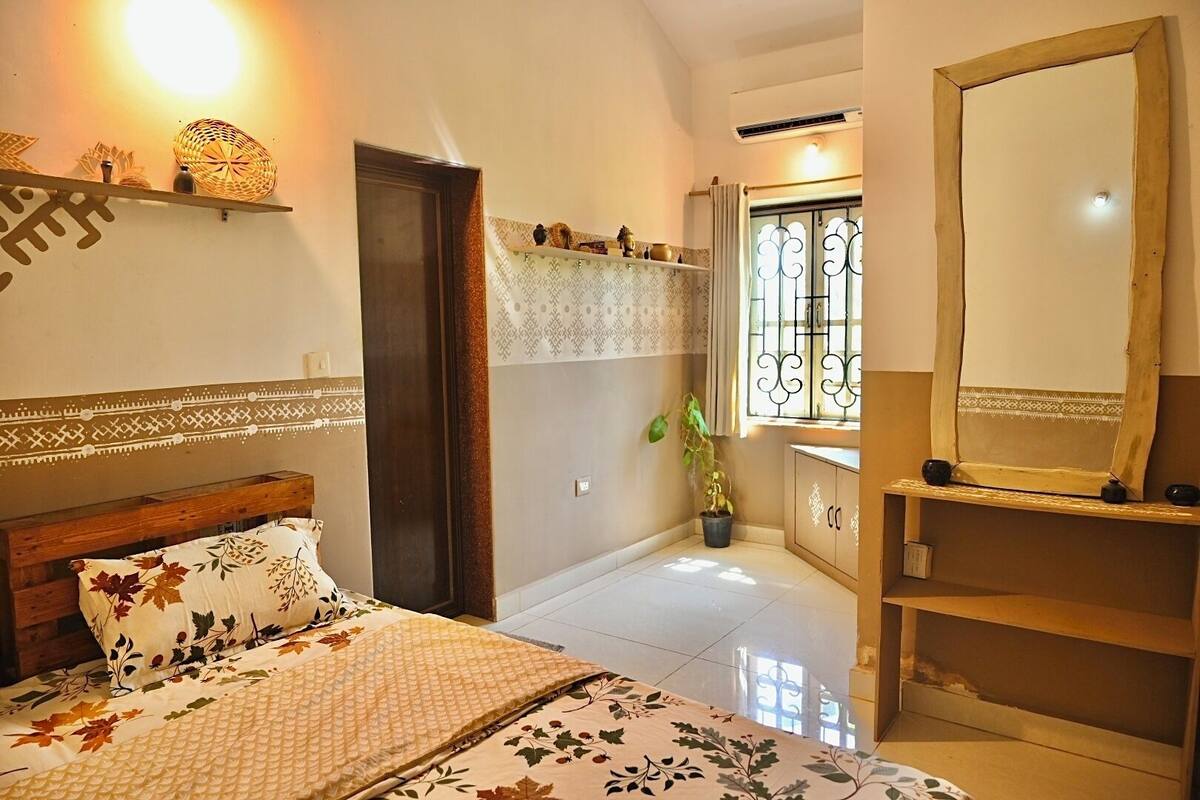 A bedroom with a bed, a bedside table, a mirror, a potted plant, and a window with a curtain.