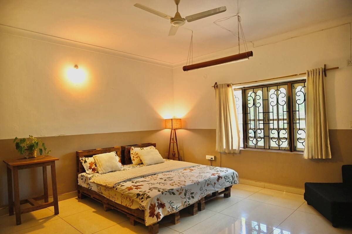 A bedroom with a bed, a standing lamp, a ceiling fan, a window with curtains, and a potted plant.