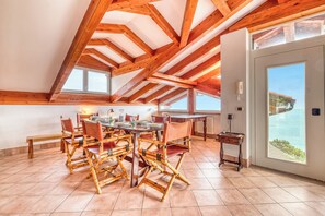 A dining area with a high ceiling and wooden beams, a large table with chairs, and a view of the sea through a large window.