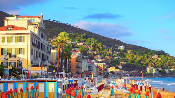 Eine Küstenstadt mit bunten Gebäuden, einem Strand mit Sonnenschirmen und einer Hügelkulisse im Hintergrund.