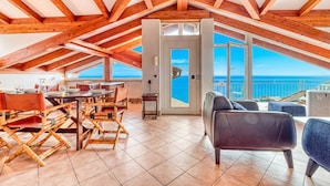 Ein Essbereich mit Holzsesseln und einem Tisch, ein Balkon mit Meerblick und eine hohe Decke mit Holzbalken.