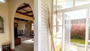 A spacious living area with a wooden floor, a dining table, and chairs. There is a large window with a view of a garden and a fence.