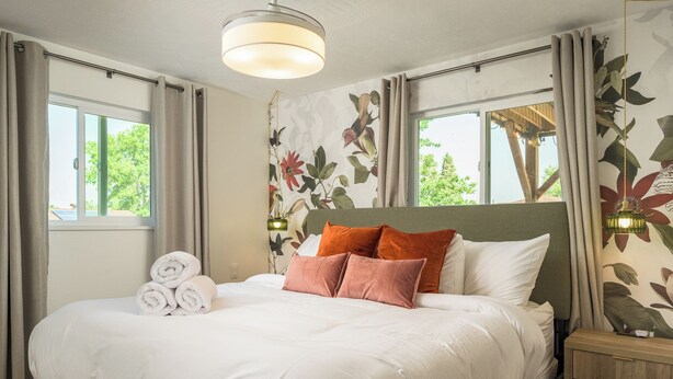 Un lit bien fait, avec du linge de lit blanc et deux coussins orange, une table de chevet en bois avec une lampe, et un papier peint à motifs floraux.
