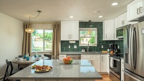 Une cuisine moderne avec des meubles blancs, un îlot central, des appareils électroménagers en acier inoxydable et une vue sur une maison par la fenêtre.