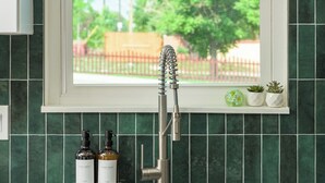 Une cuisine avec une fenêtre, des carreaux verts, un robinet et deux bouteilles posées sur le comptoir.