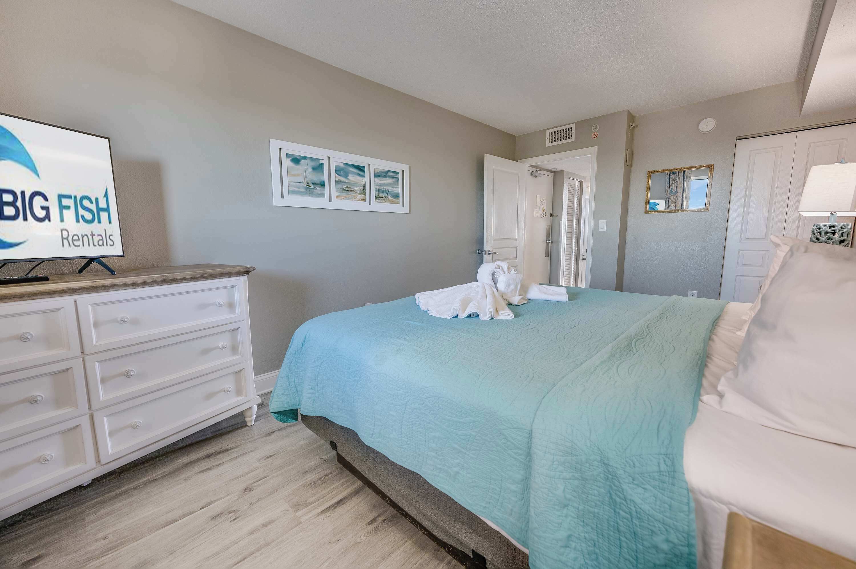 Een slaapkamer met een bed, een kaptafel, een televisiescherm waarop 'BIG FISH Rentals' staat, en drie ingelijste foto's aan de muur.