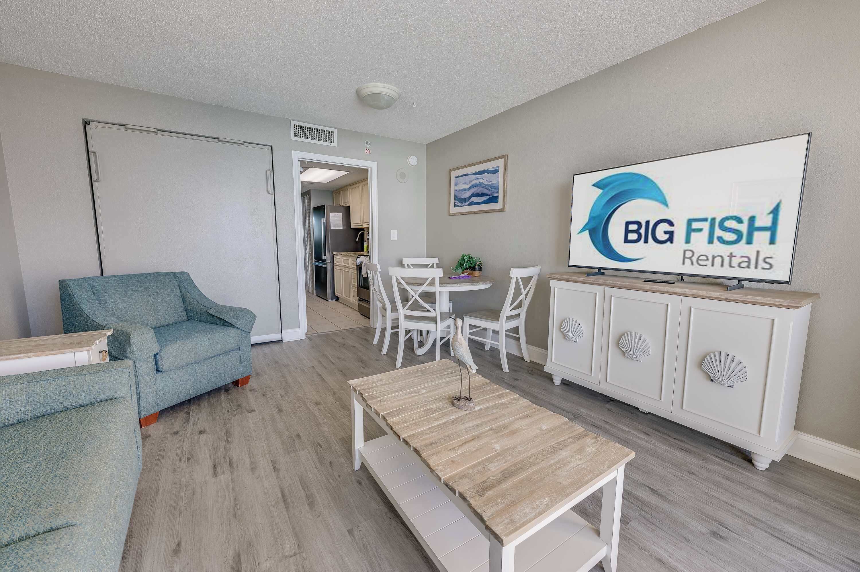 A modern living room with a sofa, a coffee table, and a dining area with chairs and a table. A TV is mounted on the wall displaying the logo "BIG FISH Rentals".