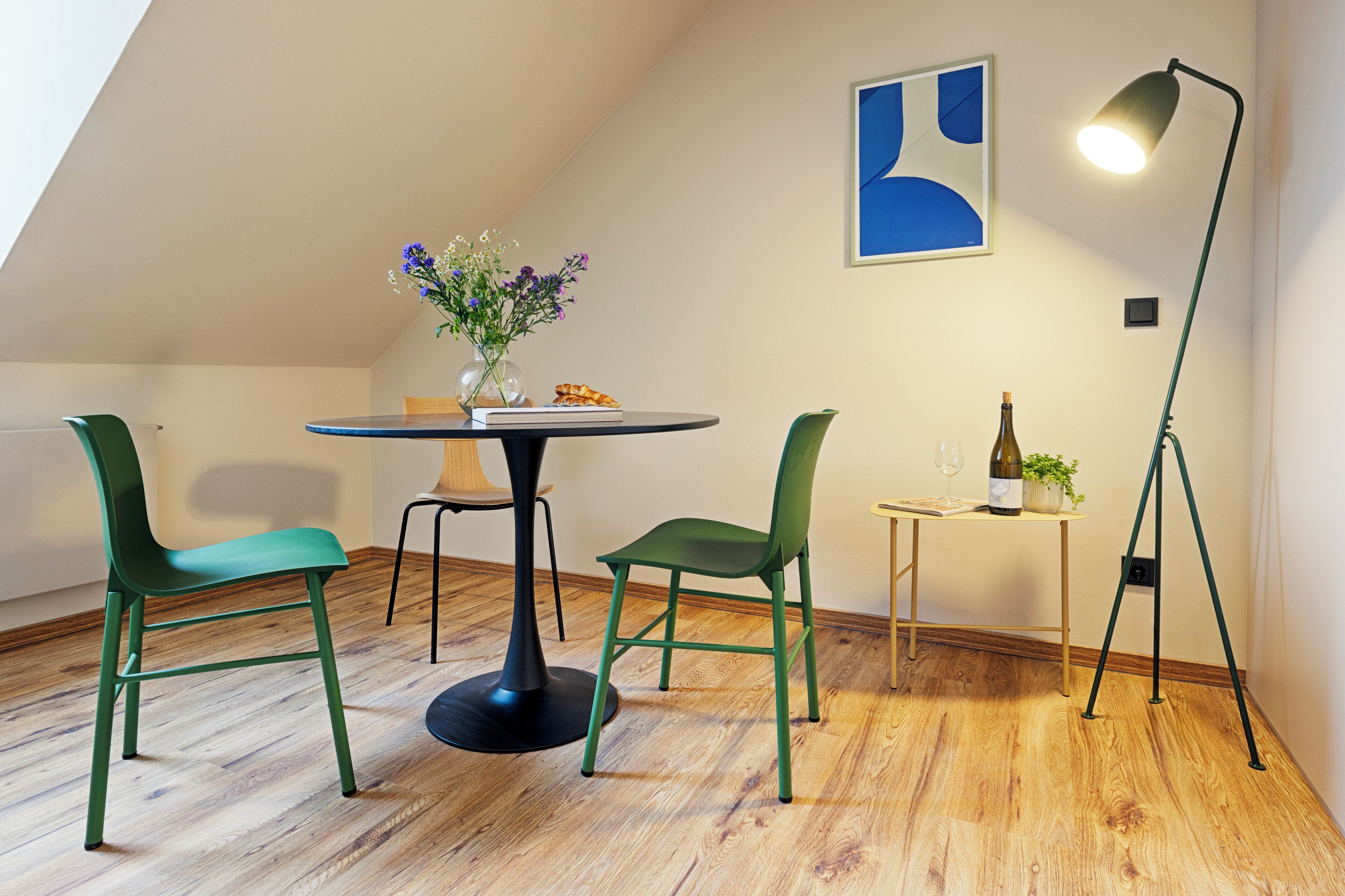 A dining area with a round table, two green chairs, a vase with flowers, a bottle of wine, and a lamp.