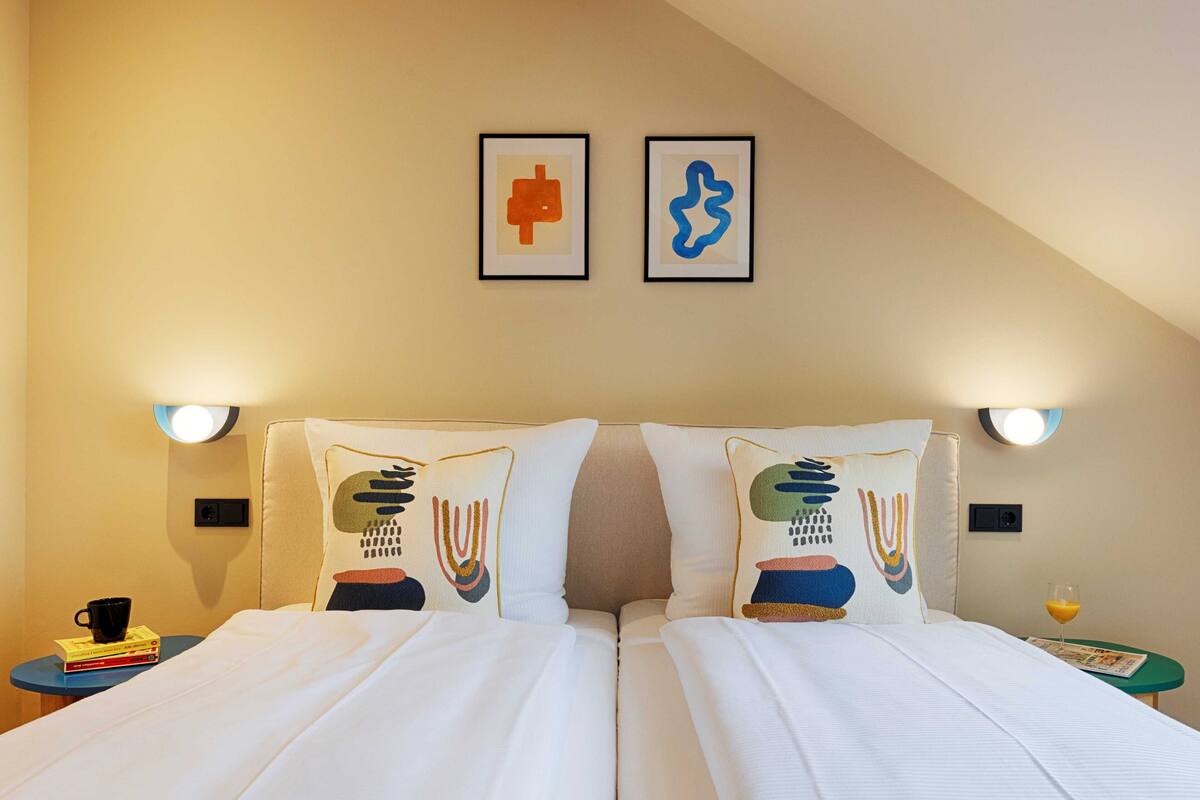 Une chambre d’hôtel avec un lit, deux oreillers aux motifs originaux, une table de chevet avec une tasse et des livres, et des œuvres d’art encadrées accrochées au mur.
