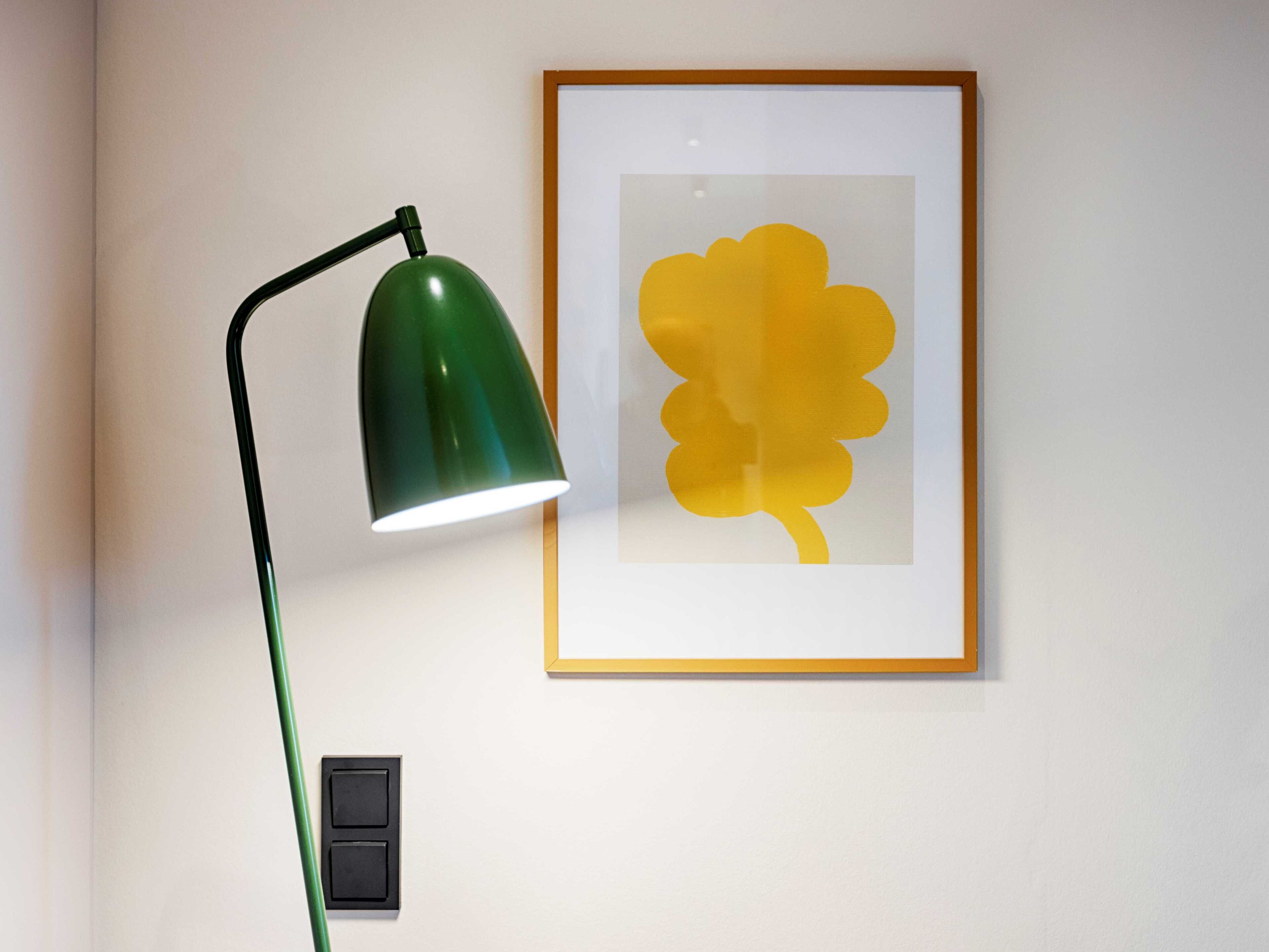 A green desk lamp with a white shade, a framed yellow artwork on the wall, and a black switch on the wall.