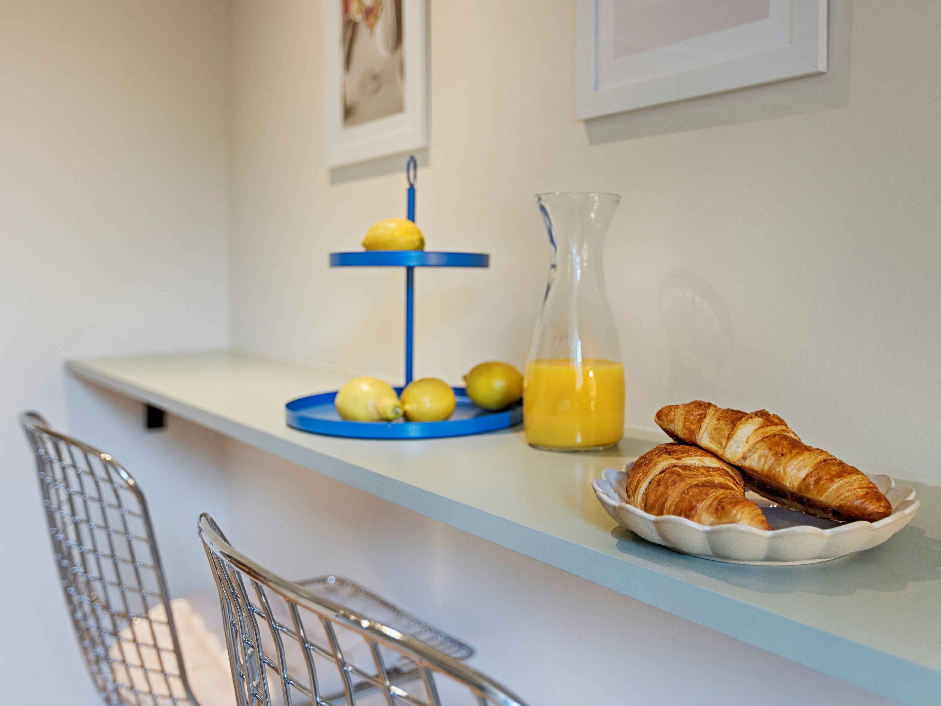 Ein Frühstücksarrangement mit einer Croissantplatte, einem Glas Orangensaft und einem blauen Obstkorb mit Zitronen.