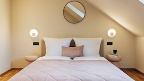 A bedroom with a large bed, two bedside tables, a round mirror on the wall, and two wall-mounted lights.