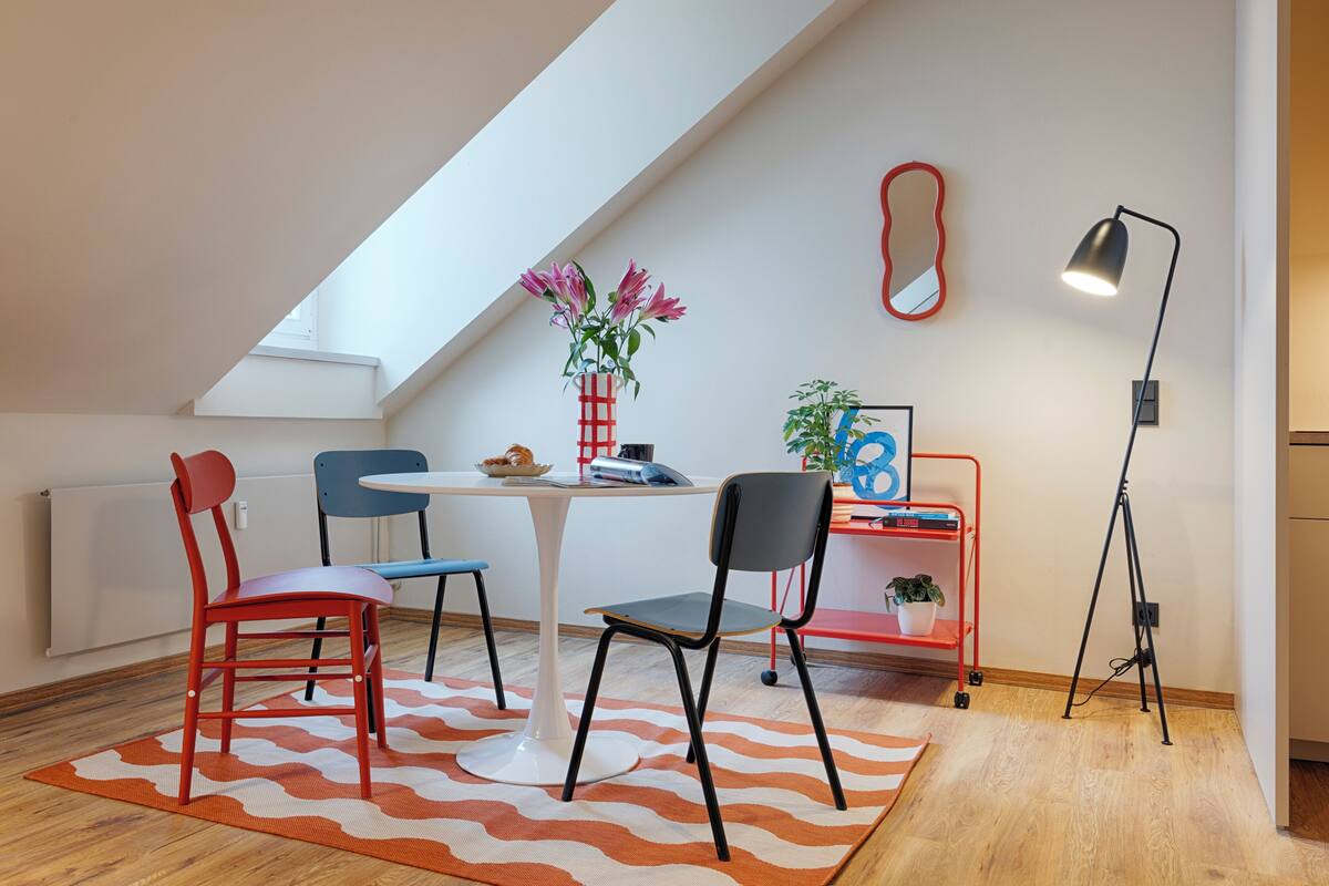 Une cuisine moderne avec un coin repas, comprenant une table blanche, des chaises rouges et bleues, un chariot à chaises rouge et bleu, une lampe sur pied et une plante en pot.