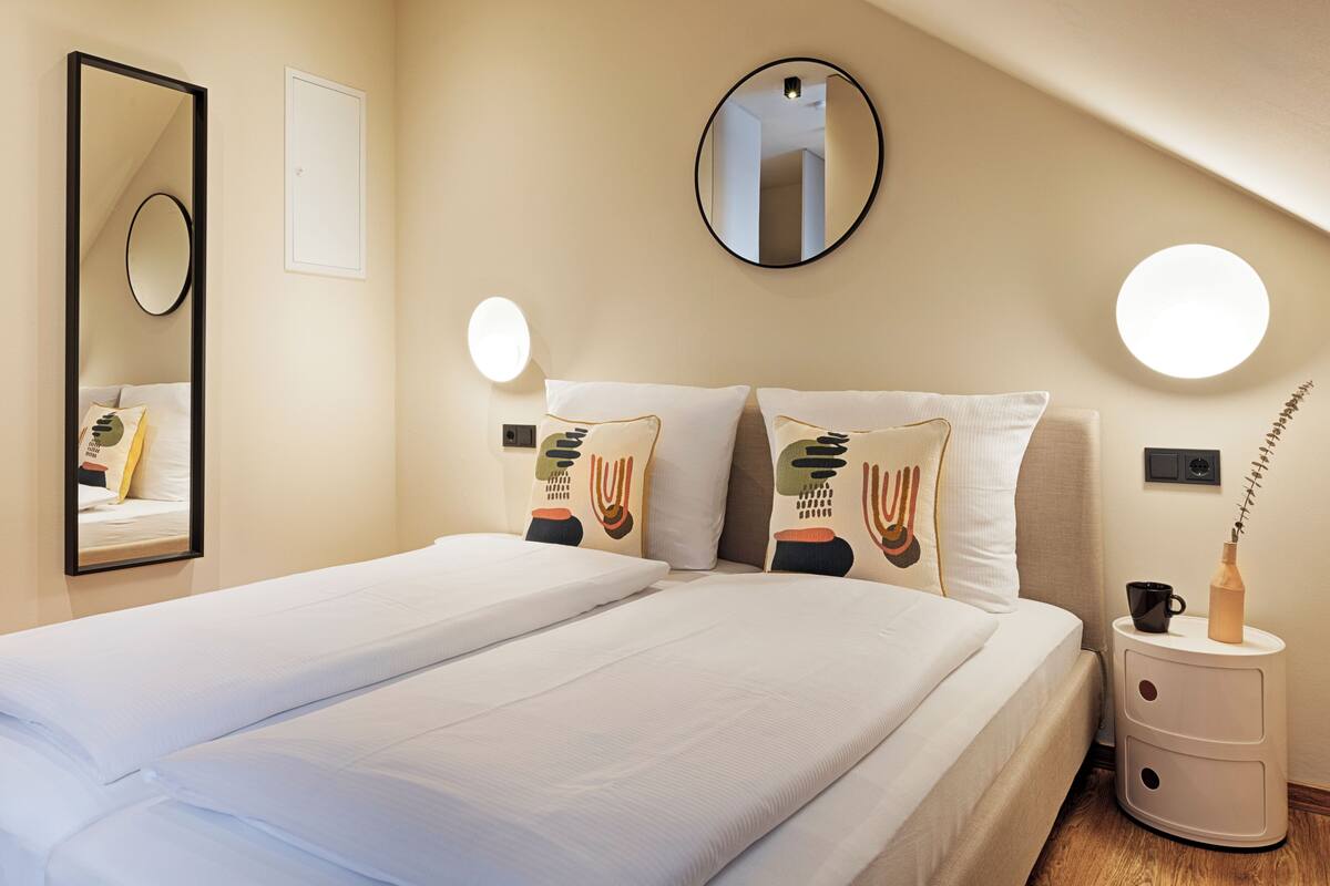 Un lit bien fait, avec du linge de lit blanc, un miroir rond et deux lampes fixées au mur.