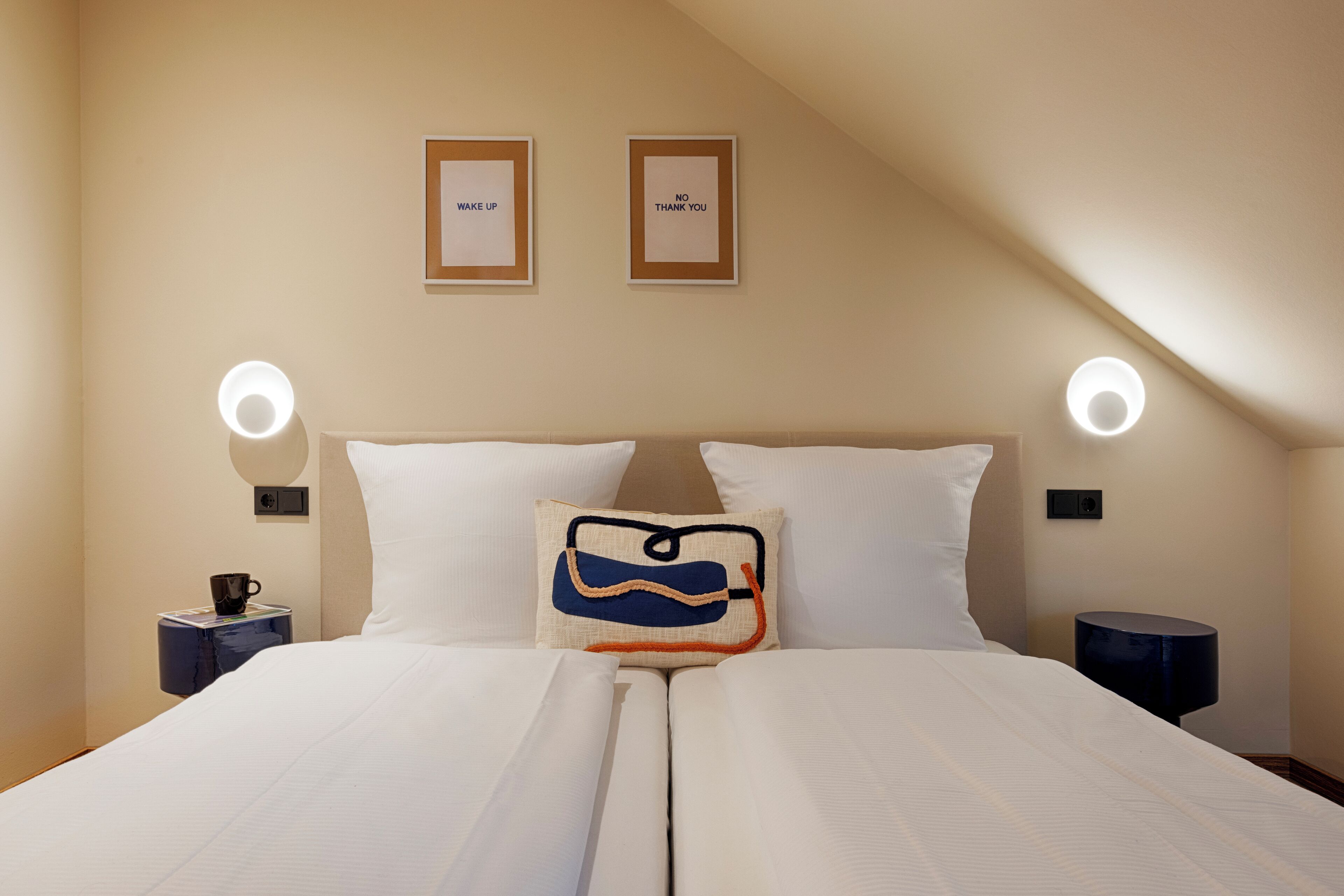 A neatly made bed with white linens, two pillows, a decorative pillow with a blue and orange design, and two wall-mounted lamps.