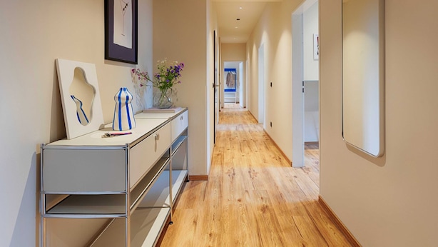 A hallway with a wooden floor, a white console table with a vase of flowers, and a framed picture on the wall.