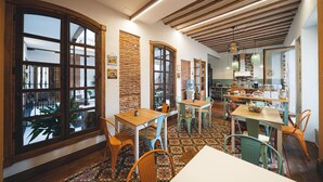 Un comedor con mesas y sillas de madera, ventanas amplias y una cocina al fondo.