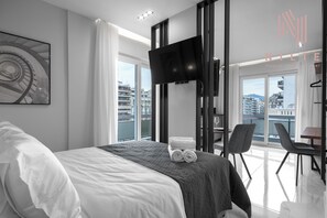 A modern hotel room with a large bed, a flat-screen TV, a balcony with a city view, and a dining area.