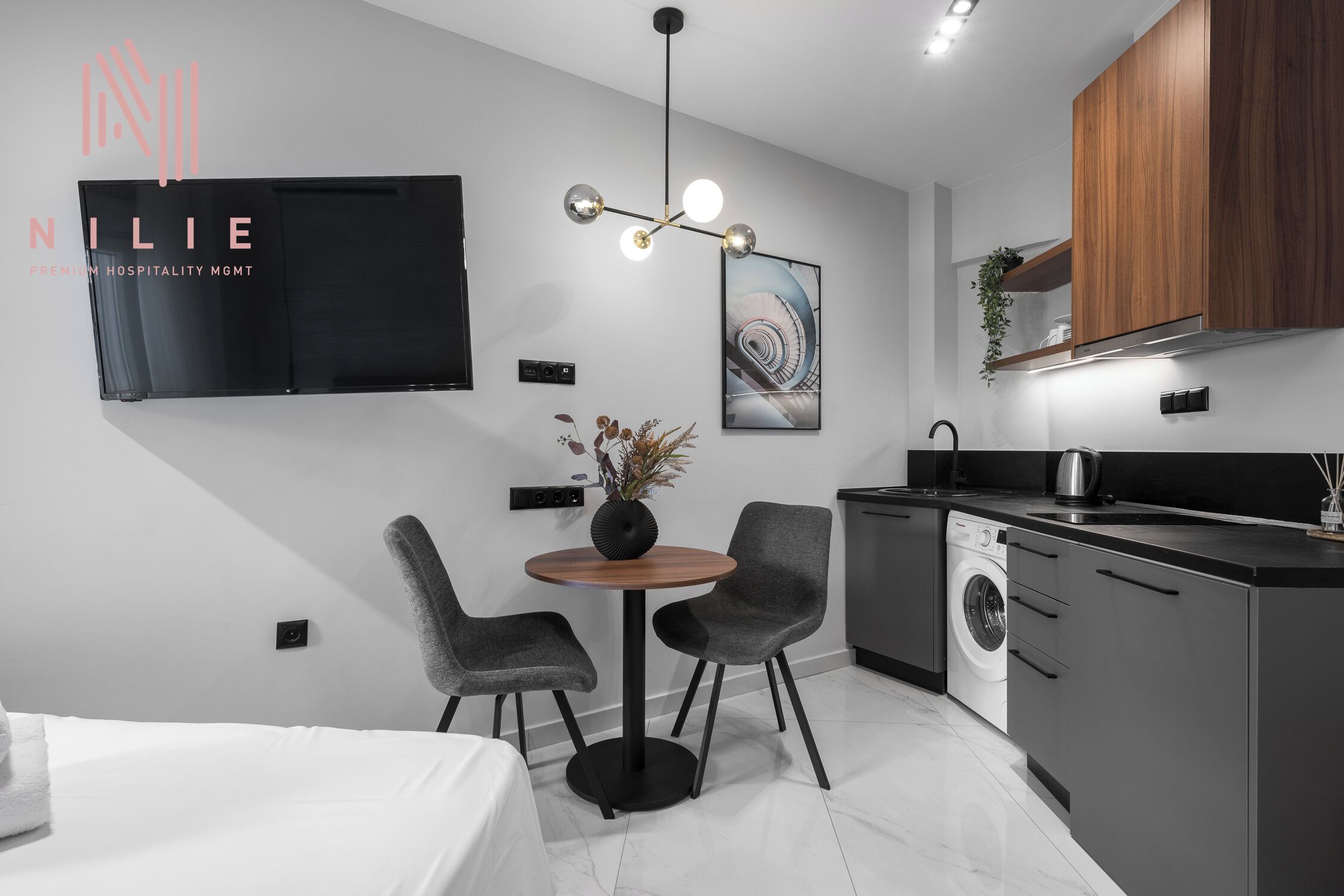 A modern hotel room with a bed, a round dining table with chairs, a TV mounted on the wall, a kitchen area with a washing machine, and a ceiling light fixture.