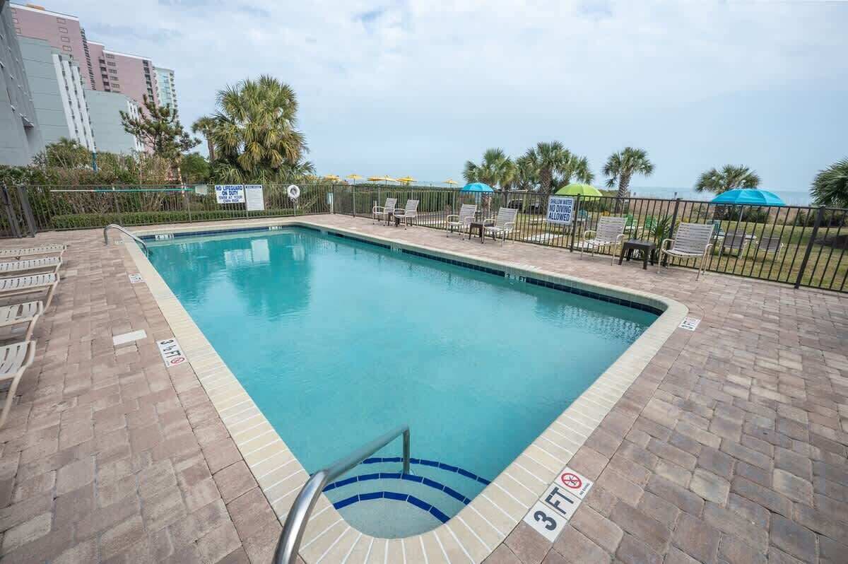 A rectangular pool with a 3ft depth marker, surrounded by lounge chairs and umbrellas, with a view of the ocean and palm trees.