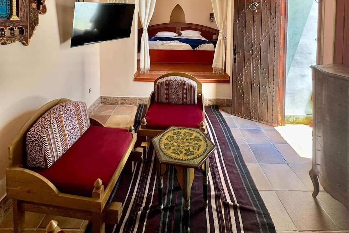 Quarto com cama, sofá, pequena mesa e televisão fixada na parede.