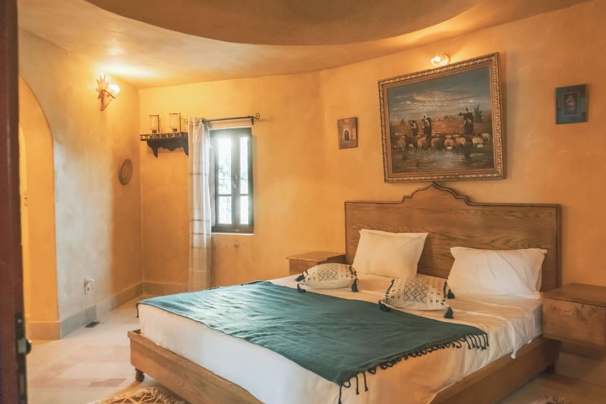Um quarto com cama de madeira, duas mesas de cabeceira, um criado-mudo, uma janela com cortinas, uma luminária na parede e um quadro emoldurado.