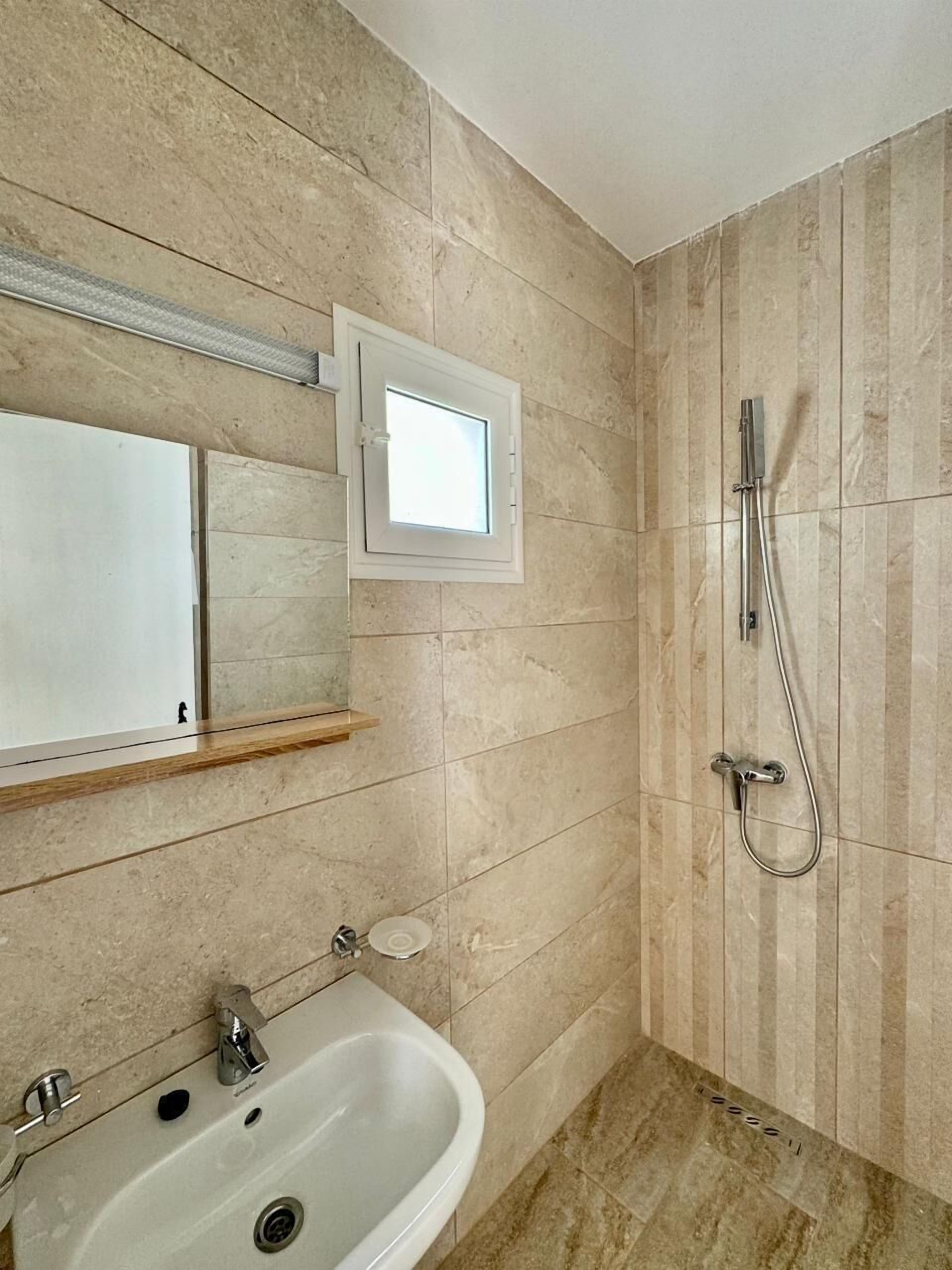 A bathroom with a sink, mirror, shower, and a small window.