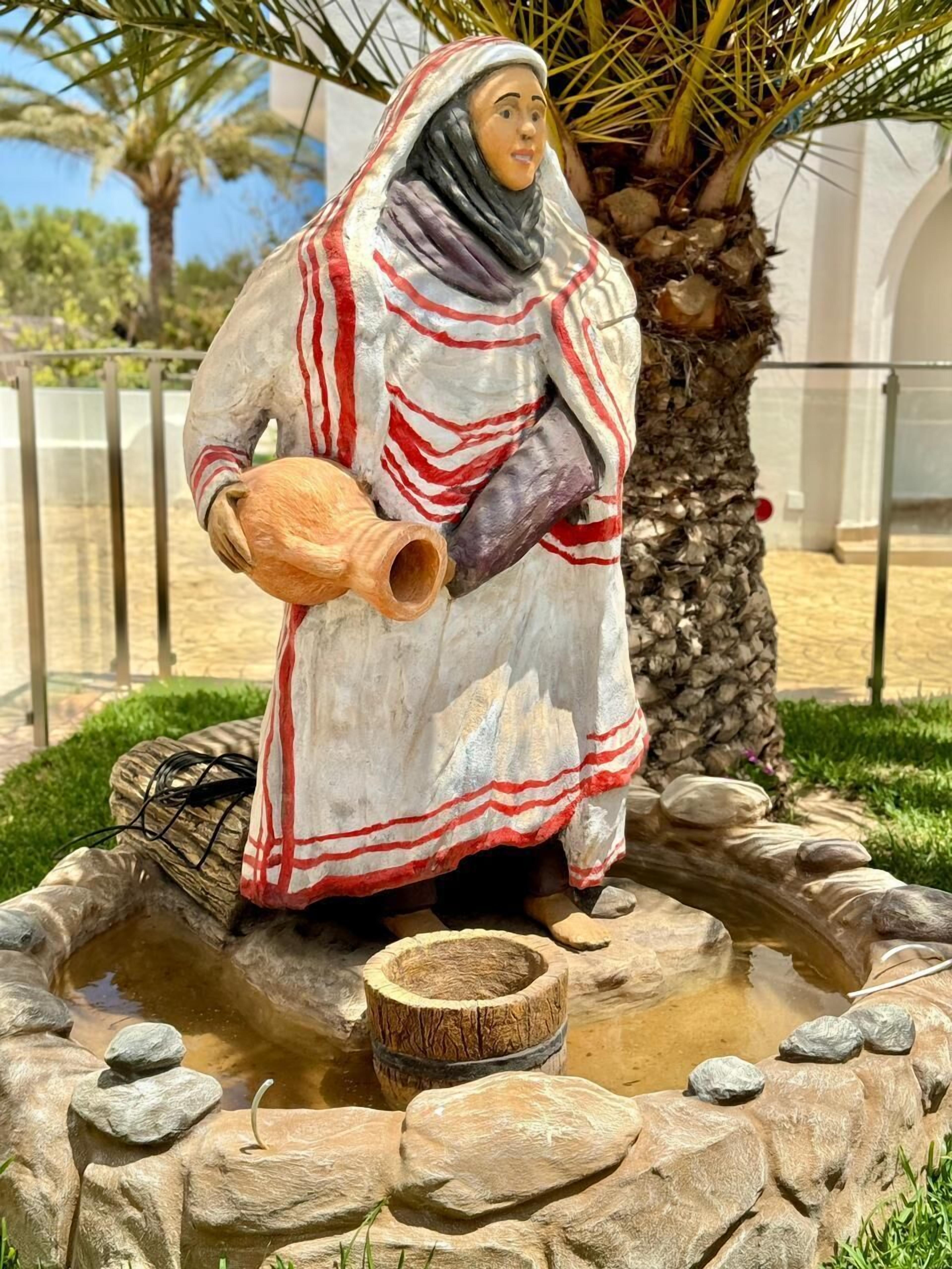 Una estatua de una persona vestida con atuendo tradicional, sosteniendo una olla de barro, ubicada en una fuente de agua rodeada de rocas y palmeras.