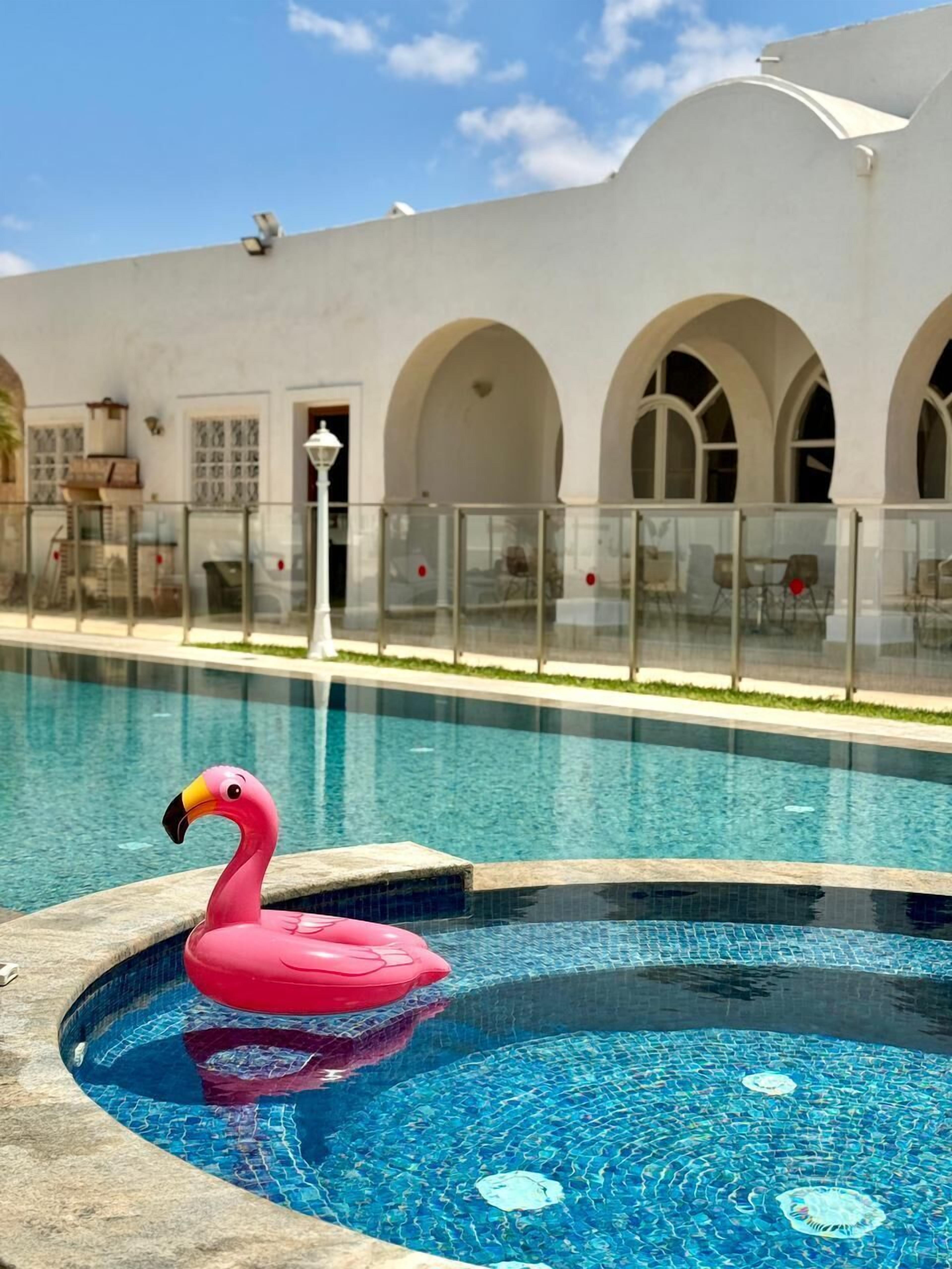Une piscine avec un flotteur en forme de flamant rose, entourée d’un bâtiment blanc aux fenêtres en arc de cercle, sous un ciel d’un bleu limpide.