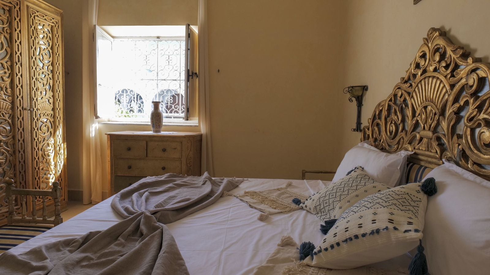 A bedroom with a bed, a wooden nightstand, a decorative headboard, and a window with a view.