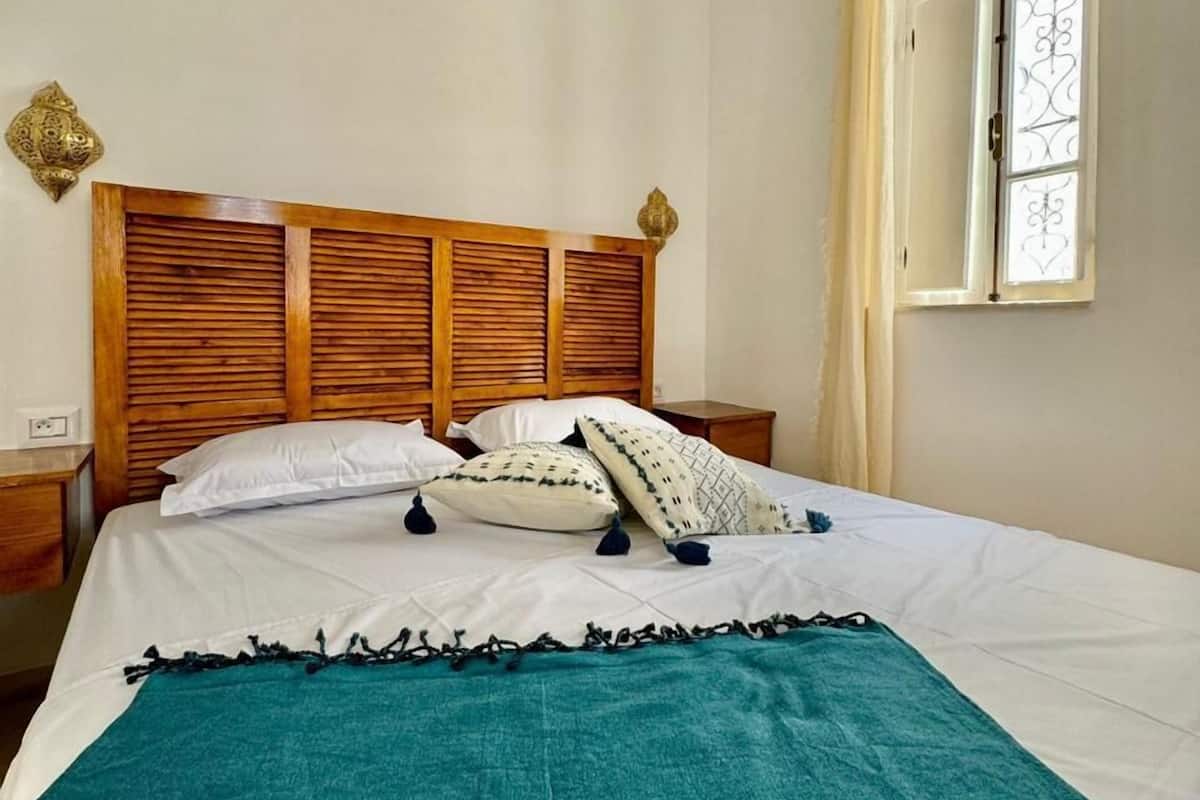 Cama bem arrumada com cabeceira de madeira, roupa de cama branca e manta azul-esverdeada.