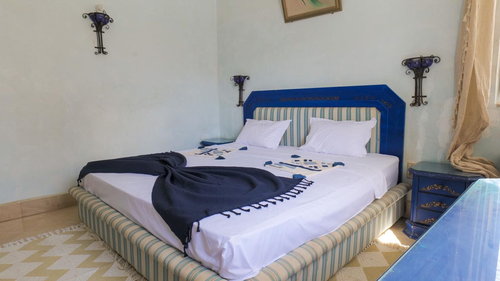 A bed with a striped headboard, white bedding, and a dark throw blanket. There are two wall-mounted lamps and a small bedside table with a lamp.