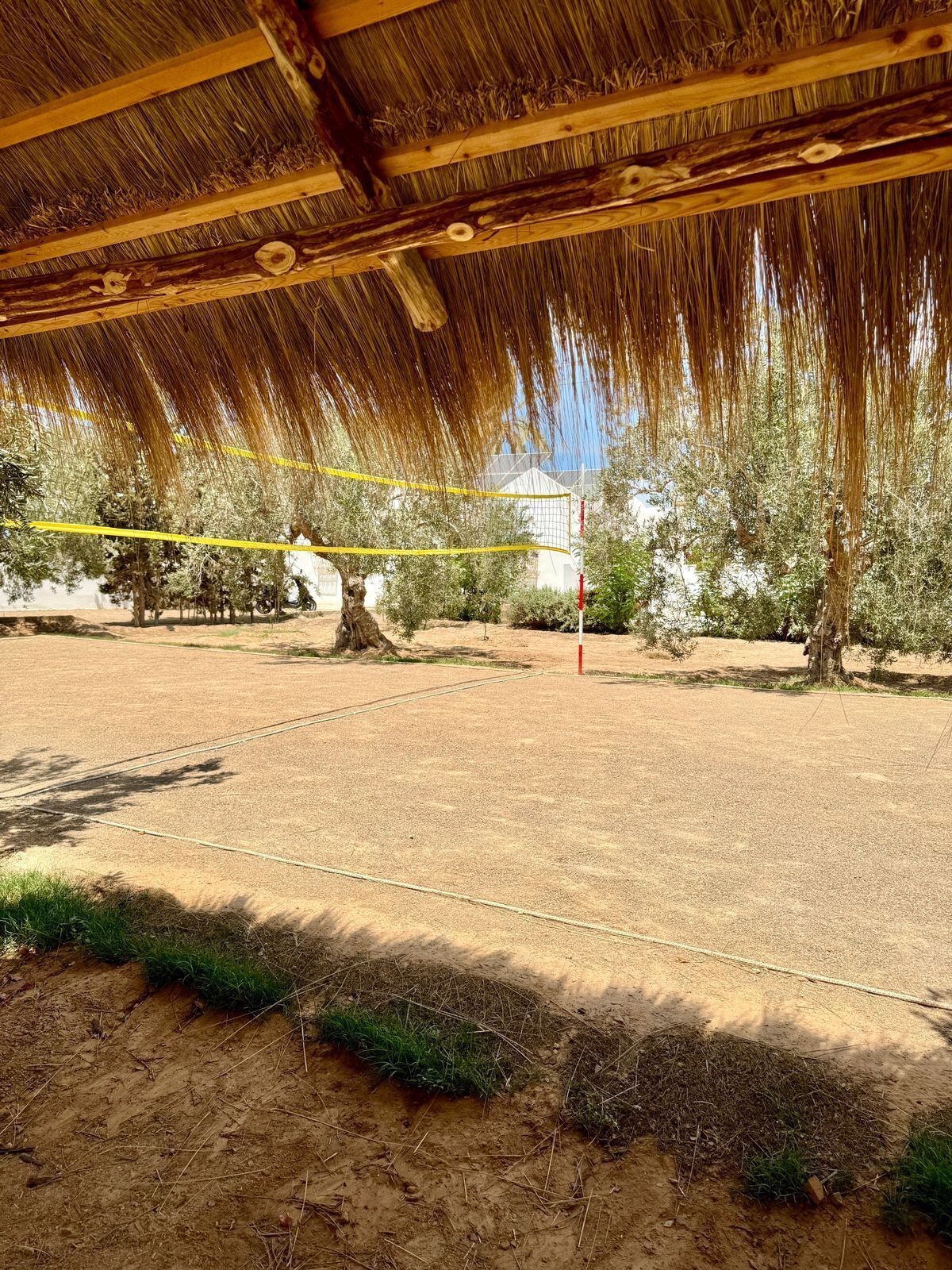 Un terrain de sport intérieur, doté d’un filet, entouré d’arbres et surmonté d’un toit de chaume.
