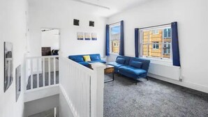 Un salon moderne avec un canapé bleu, des murs blancs et une grande fenêtre donnant sur des immeubles.