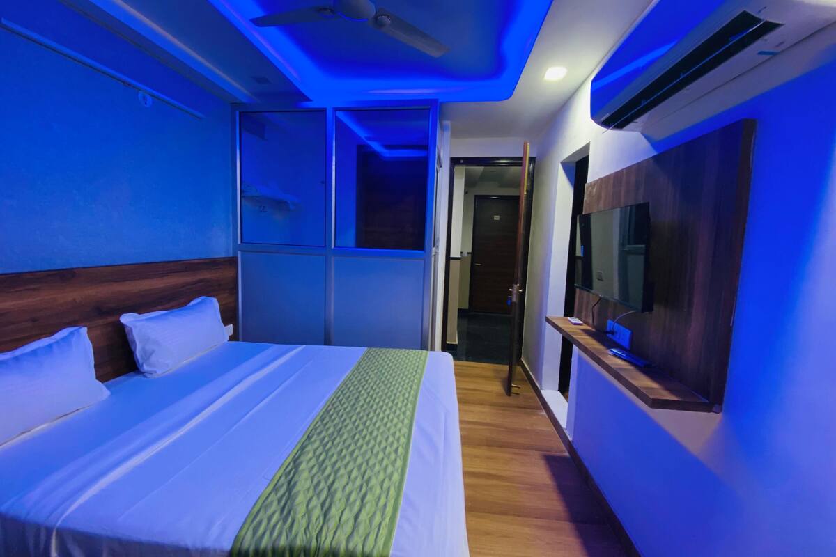 A hotel room with a bed, a ceiling fan, an air conditioning unit, a television, and a door leading to another room.