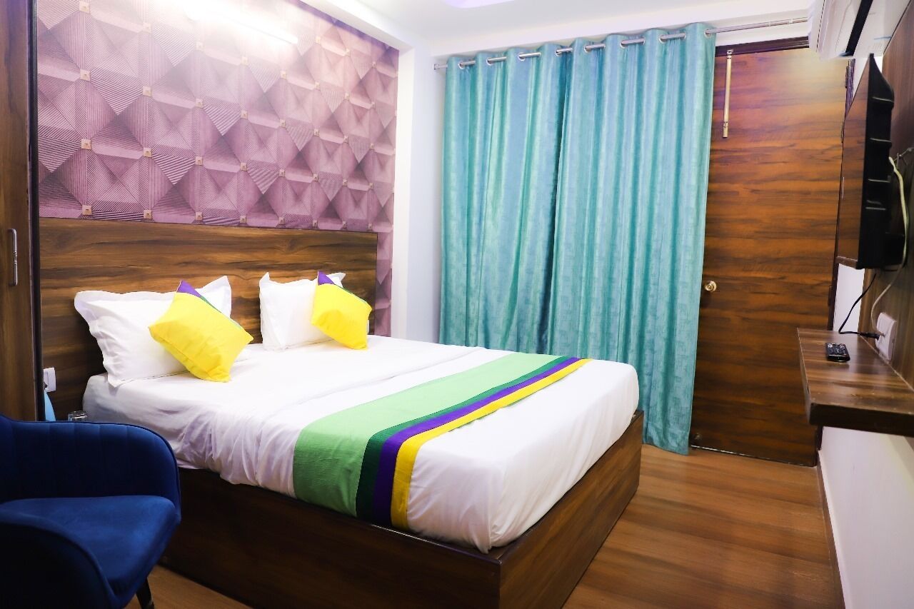 A hotel room with a bed, a blue chair, a wooden headboard, a TV, and a curtain.