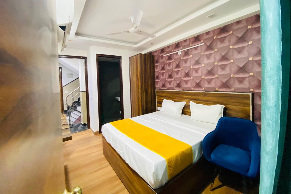 A hotel room with a bed, a blue chair, a ceiling fan, and a staircase in the background.