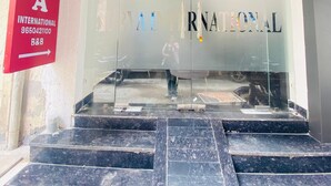 A building entrance with a glass facade, a red sign on the left, and a person standing in the doorway. - Hotel Surya International (New Delhi)