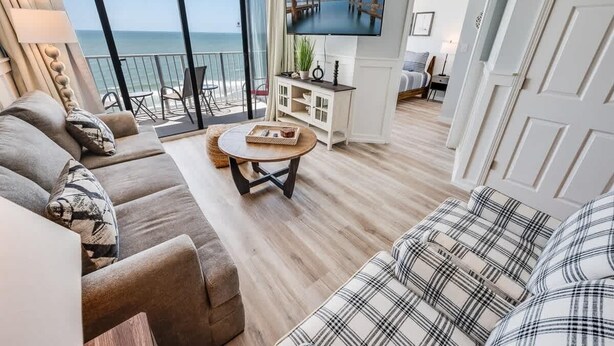 A living room with a sofa, a coffee table, and a TV. There is a balcony with a view of the sea. - Direct Ocean View - Exclusive Sand Beach Club 2BR (Myrtle Beach)