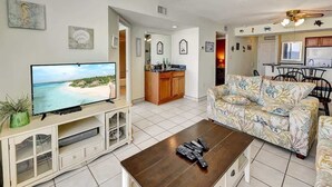 A living room with a TV, a coffee table, a sofa, and a kitchen area in the background. - New, Low Floor, Direct Ocean View Sands Beach Club (Myrtle Beach)