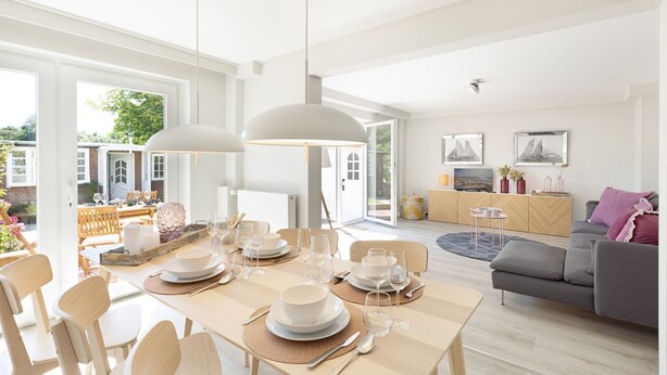 A modern dining area with a large table set for six, surrounded by wooden chairs. There is a sofa and a small side table in the background. - "Experience luxury and relaxation at the Buhne 44 vacation home on Sylt!" (List auf Sylt)