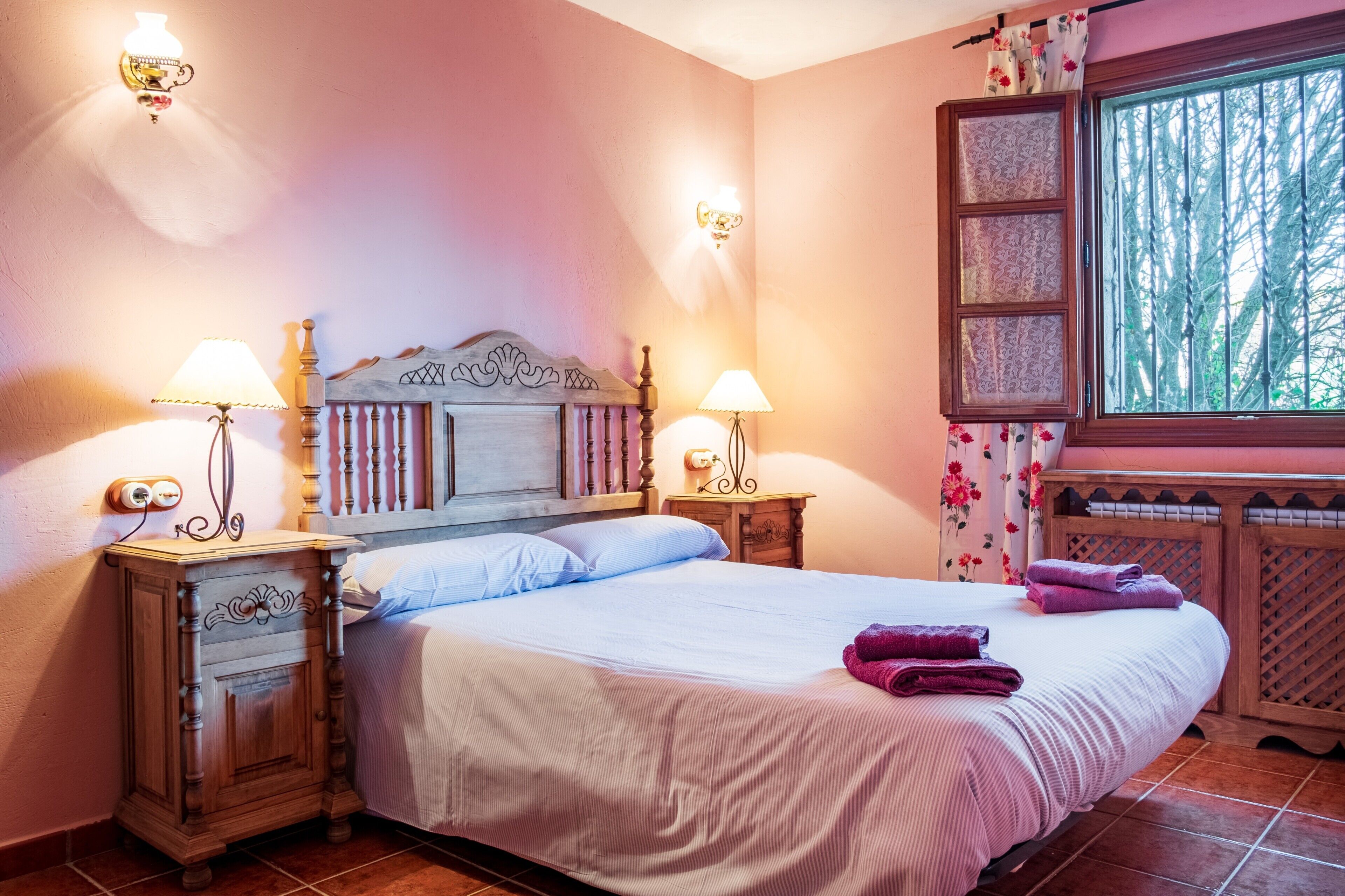 A bedroom with a wooden headboard, two bedside tables with lamps, a large bed with a white bedspread, and a window with a view of trees.