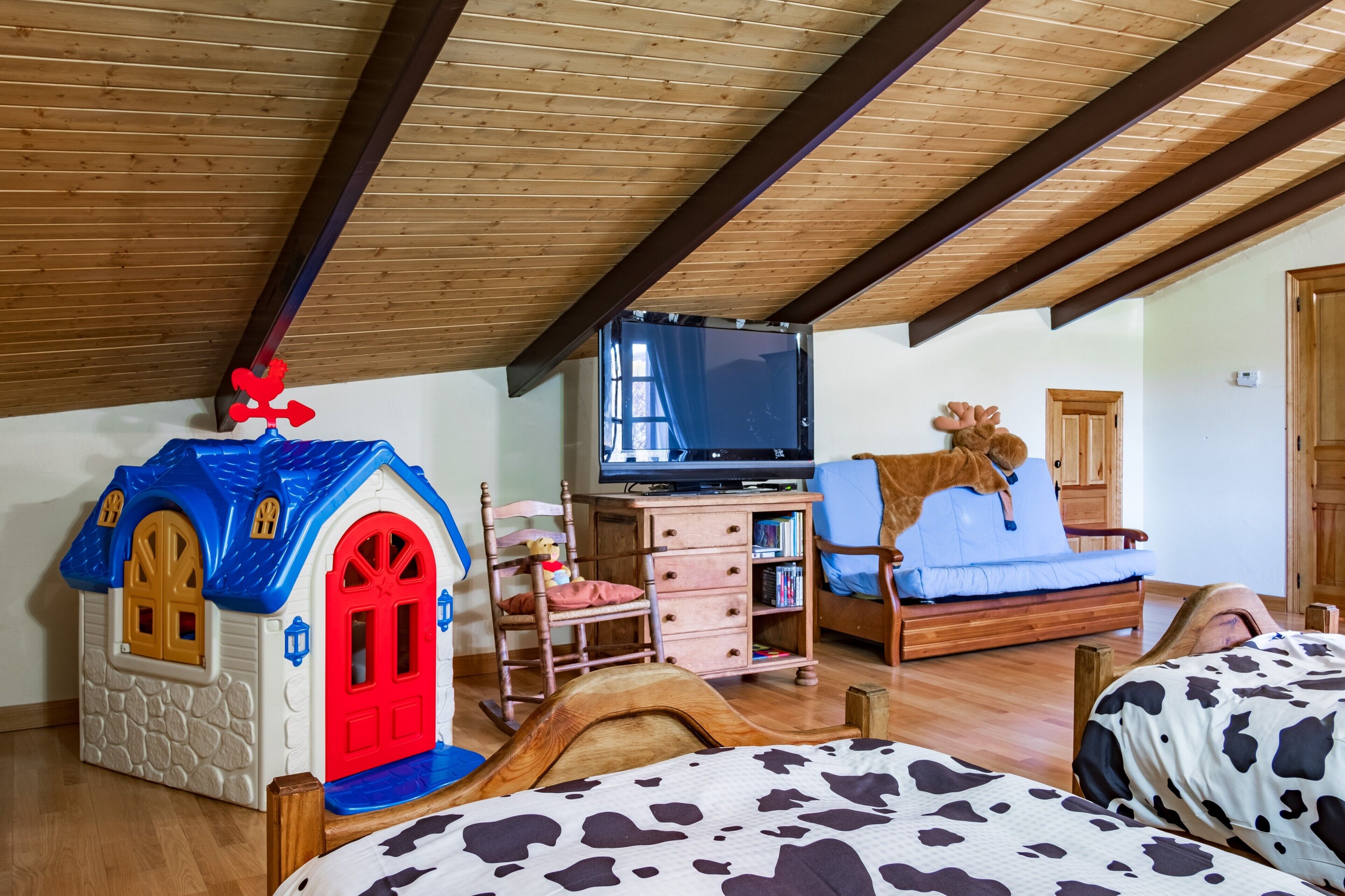 Un dormitorio con techo inclinado, televisor, cama, una casita de juegos pequeña y una silla de madera.