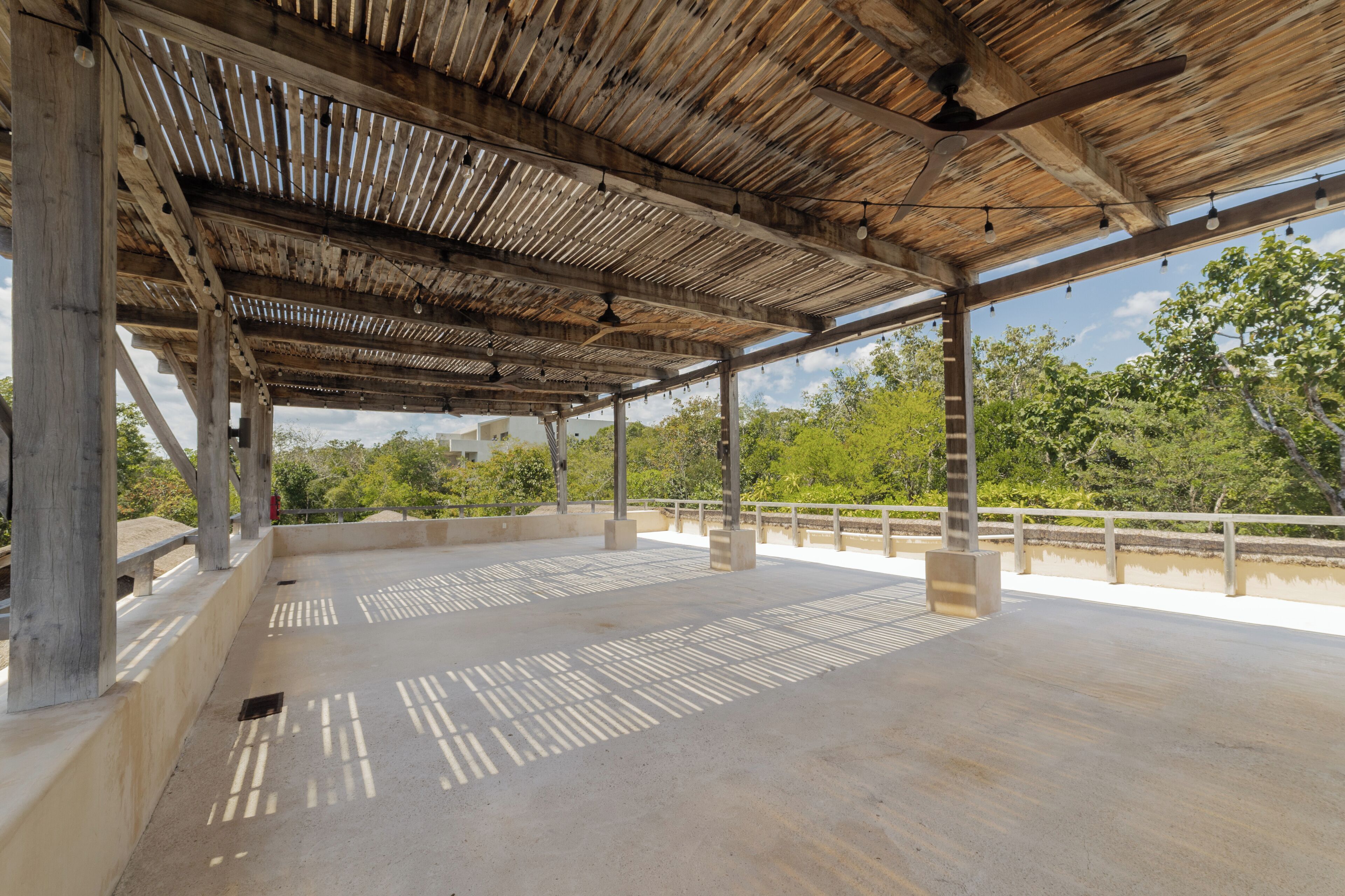 An open-air pavilion with a wooden roof and concrete floor, surrounded by greenery.