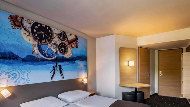 Una habitación de hotel moderna con un gran mural en la pared de relojes, una cama con sábanas blancas, una mesita de noche pequeña y un buró.