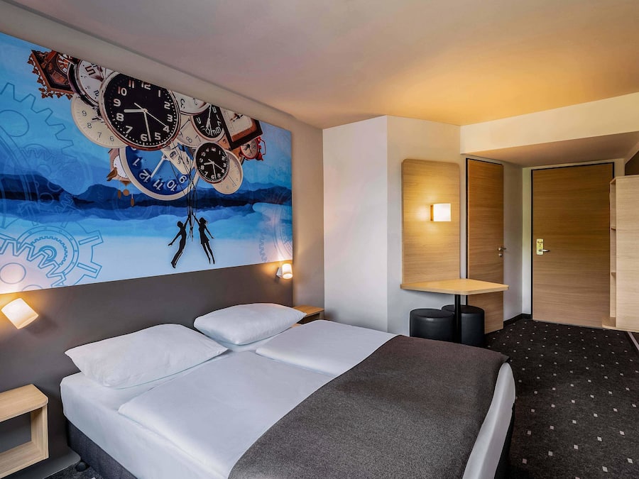 A modern hotel room with a large wall mural of clocks, a bed with white linens, a small bedside table, and a nightstand.