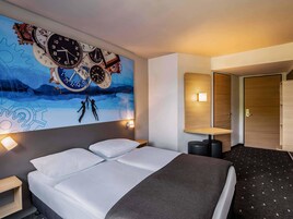 Una habitación de hotel moderna con un gran mural en la pared de relojes, una cama con sábanas blancas, una mesita de noche pequeña y un buró.